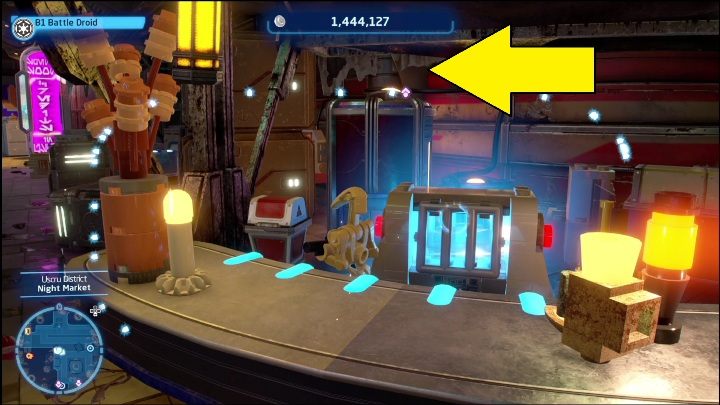 Ten klocek zamknięty został w skrzyni, aby się do niej dostać musicie wejść na teren stoiska, a następnie iść wzdłuż ściany - LEGO Skywalker Saga: Dzielnica Uscru - lista wszystkich łamigłówek - LEGO Skywalker Saga - poradnik do gry